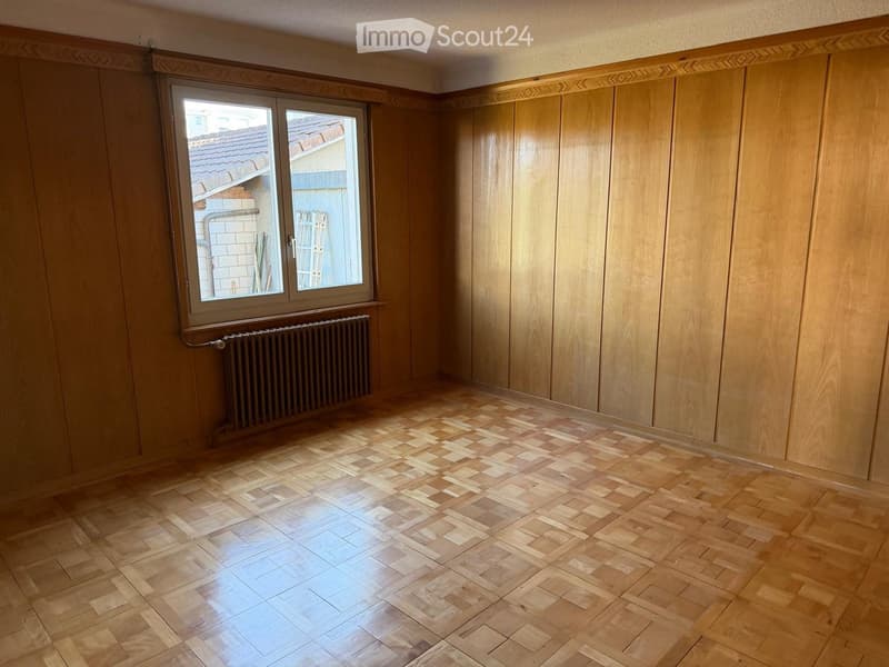 Empty room with wooden walls, herringbone parquet flooring, and a window with radiator below.