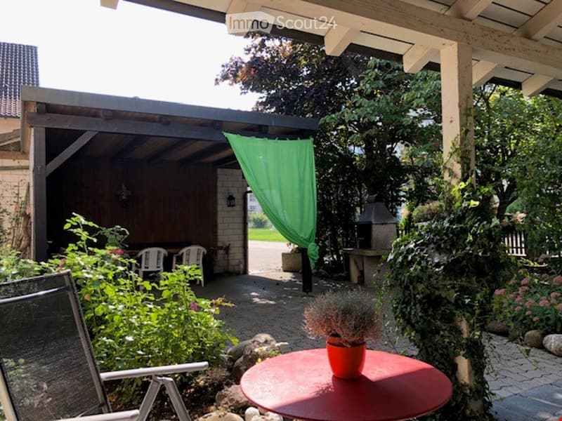 A covered outdoor patio area with a red round table, white plastic chairs, a green curtain, and surrounding greenery.