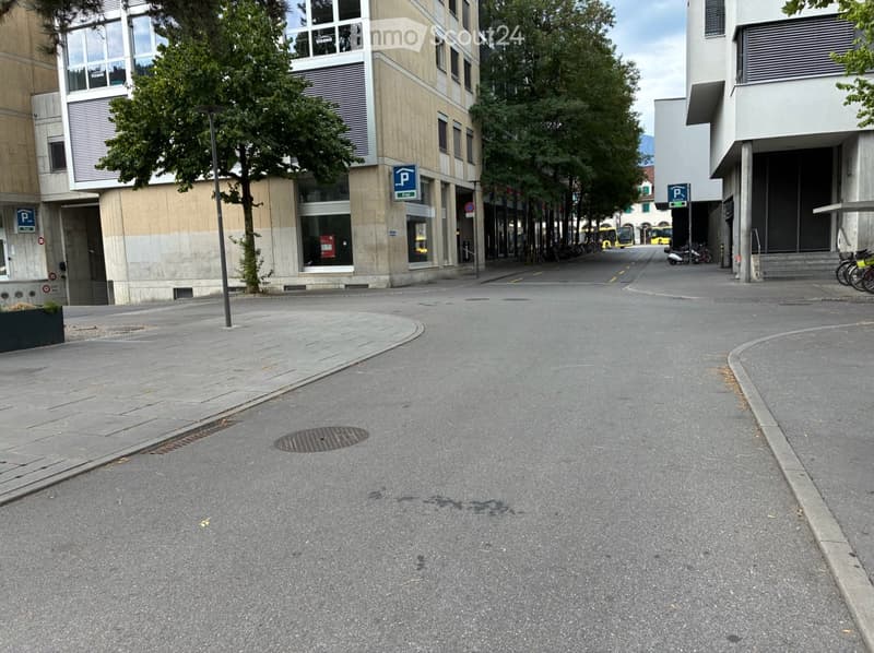 Urban street scene with modern buildings, trees, and parking signs. The road is empty, and there are no visible vehicles or pedestrians.