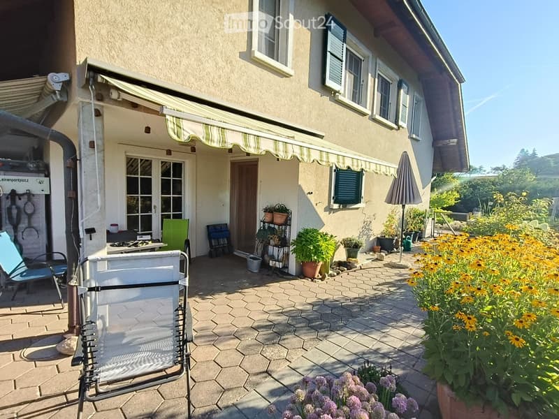 A sunny patio area with a tiled floor, outdoor furniture including chairs and a table, potted plants, and a closed umbrella. The house has a covered entrance with a striped awning and multiple windows.