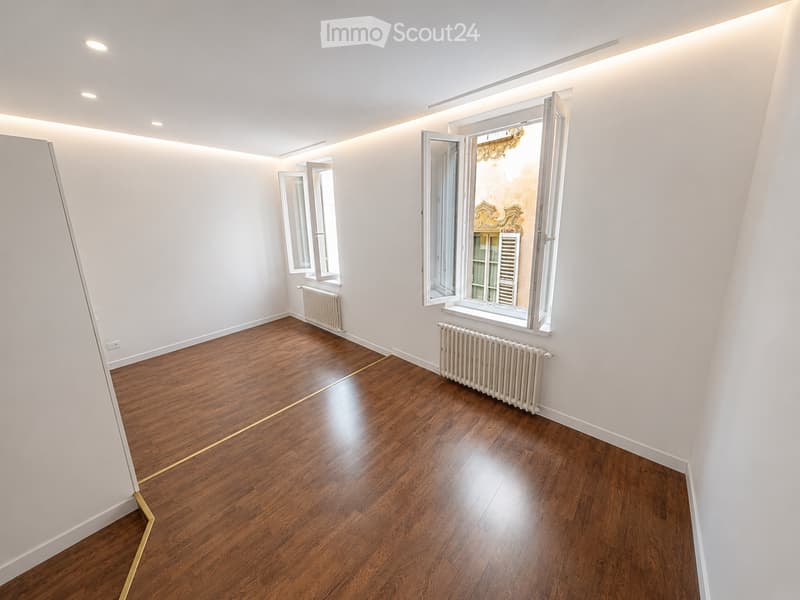 Empty room with wooden flooring, white walls, and large windows with radiators beneath them.