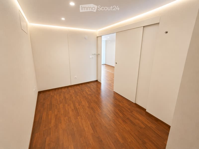 Modern hallway with wooden flooring, white walls, and recessed lighting. Features a sliding door and an open doorway leading to another room.