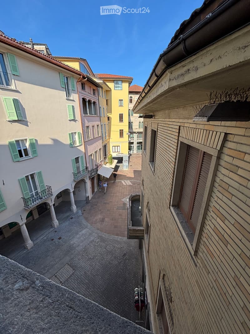 A narrow, cobblestone alleyway lined with colorful buildings featuring green shutters and balconies. The area is paved with bricks and has a few pedestrians visible.