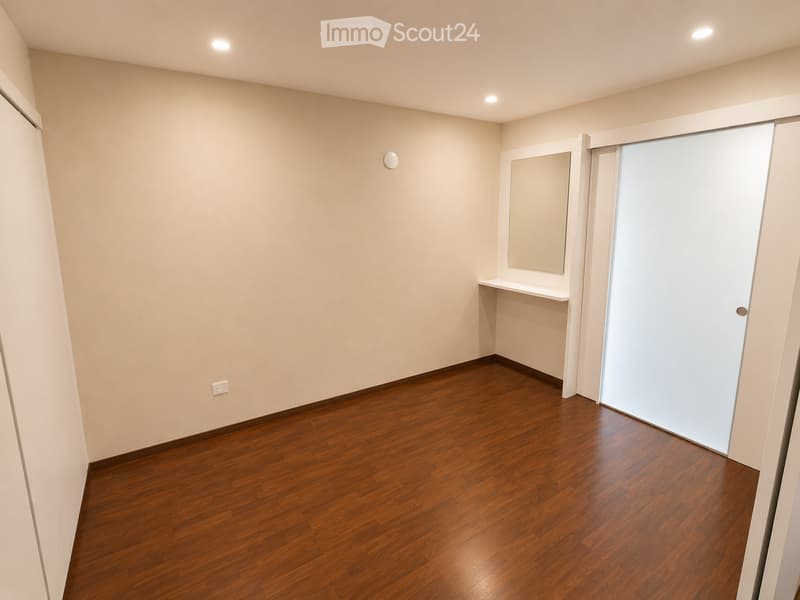 Empty room with wooden flooring, white walls, and a sliding door leading to another space. A small shelf and mirror are visible in one corner.