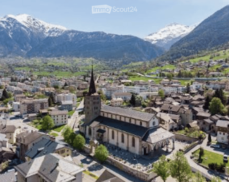 Aerial view of a European town nestled in a valley with snow-capped mountains in the background. The town features a prominent church with a tall spire, surrounded by buildings and greenery.