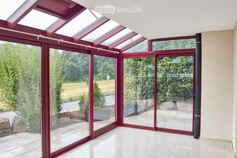 Bright, enclosed patio with large glass doors and a red-framed roof, offering views of a green landscape and garden area.