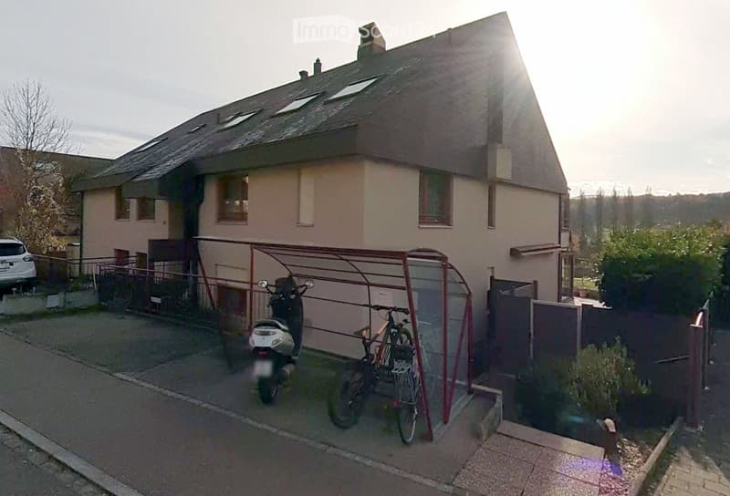 Two-story house with beige exterior, multiple windows, and a small garage. A covered parking area holds a scooter and a bicycle.