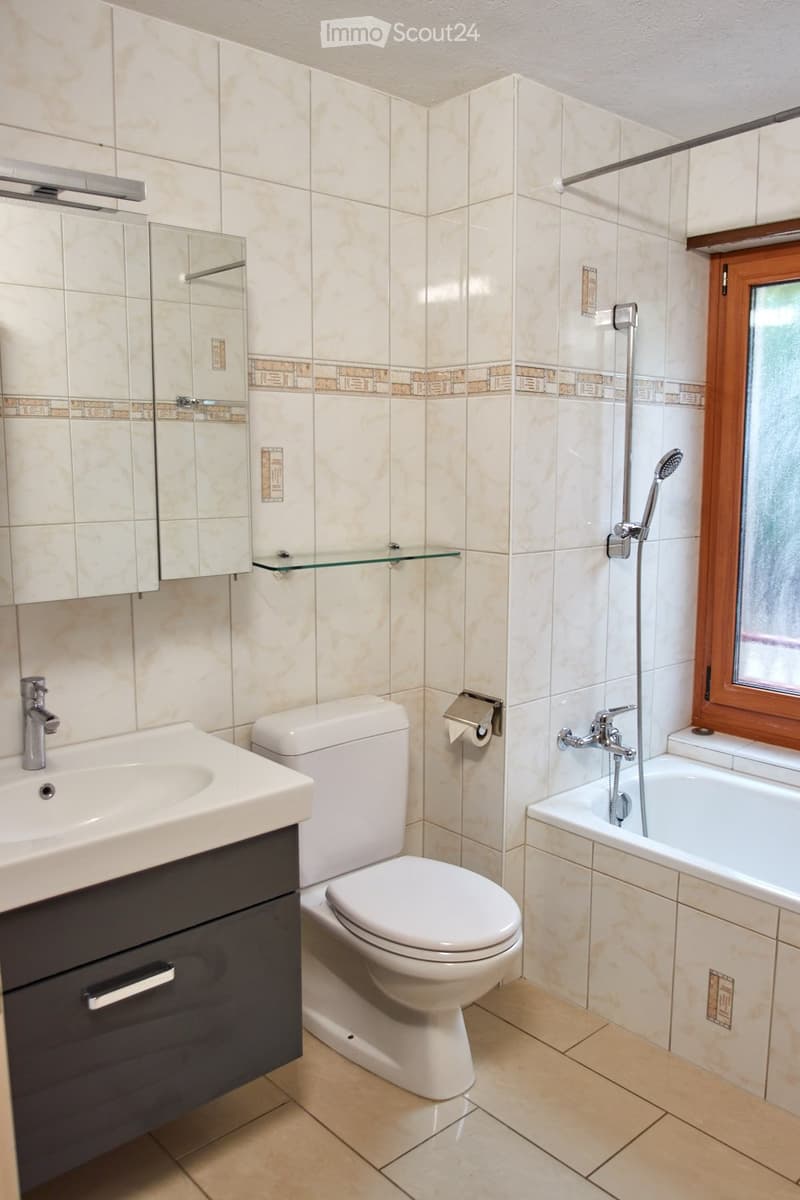 Modern bathroom with beige tiles, a white sink with a chrome faucet, a toilet, a bathtub with a handheld shower, a mirrored cabinet, and a glass shelf above the sink.