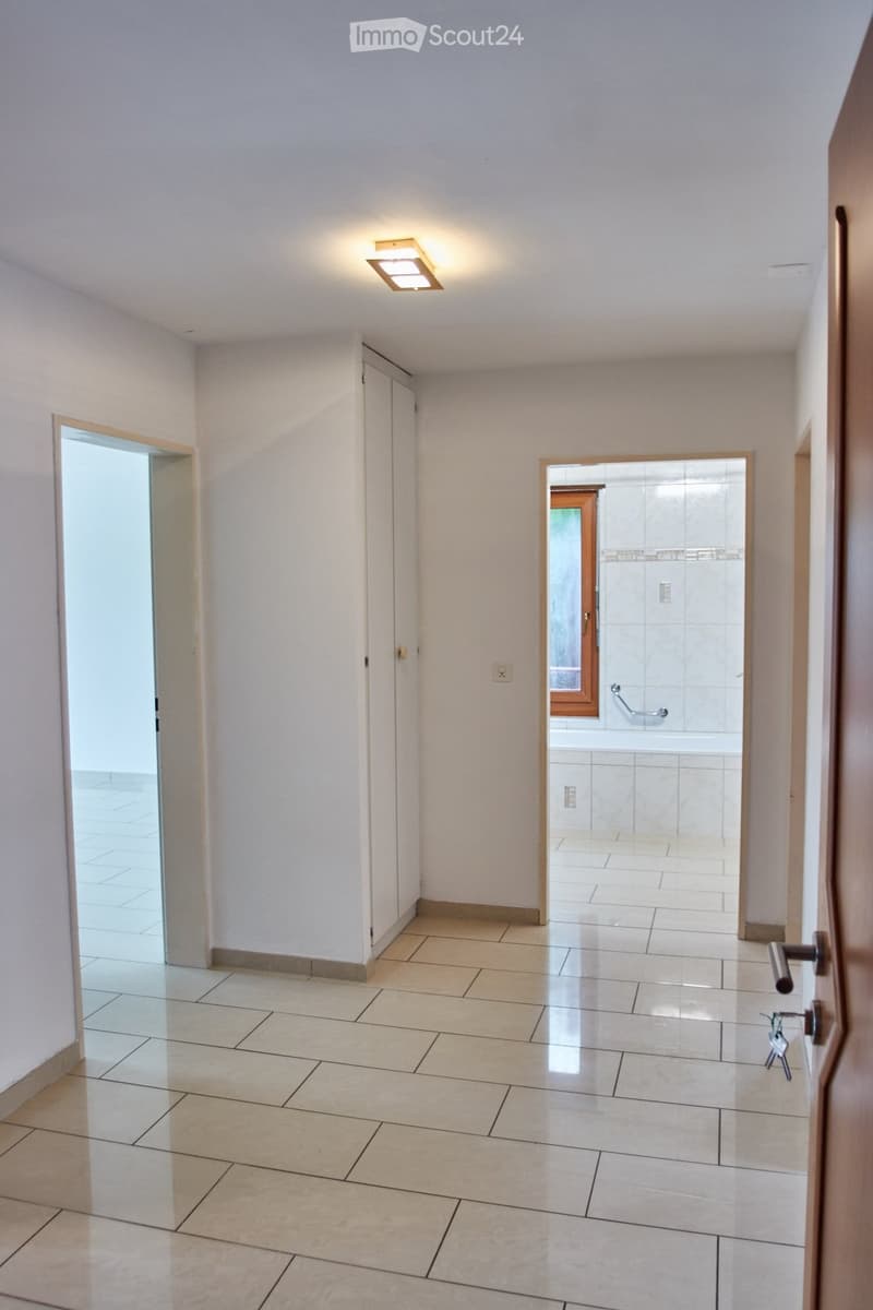 Empty hallway with tiled floor, white walls, and recessed lighting. Doors lead to adjacent rooms, including a bathroom with visible tiles and fixtures.