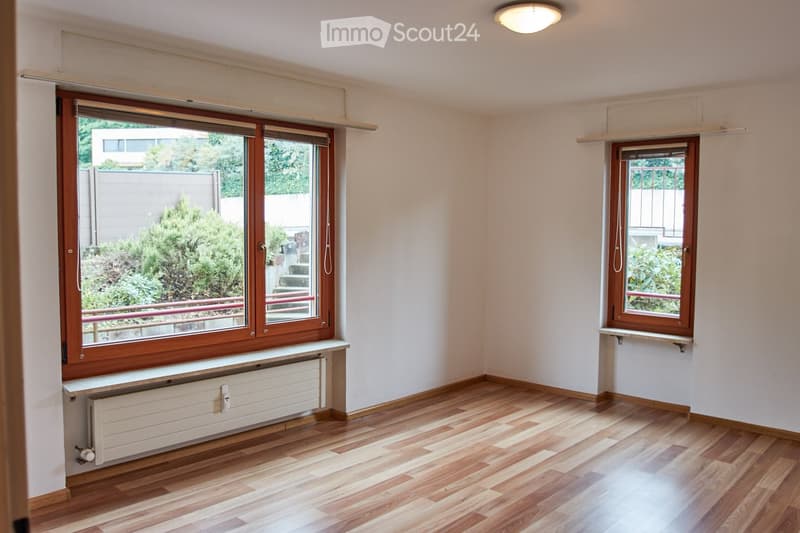 Empty room with wooden flooring, white walls, large windows with wooden frames, radiator, and a small shelf under one window.