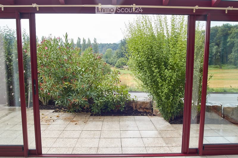 A large sliding glass door opens to a tiled balcony area with lush greenery and a view of a pond and open field beyond.
