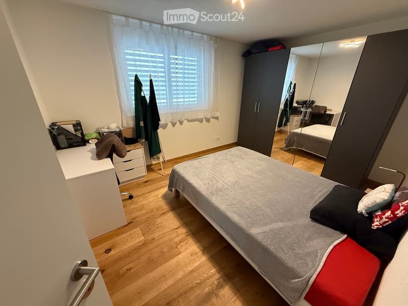 Bedroom with a double bed, mirror, wardrobe, desk, and chair. The room features wooden flooring and large windows with blinds.