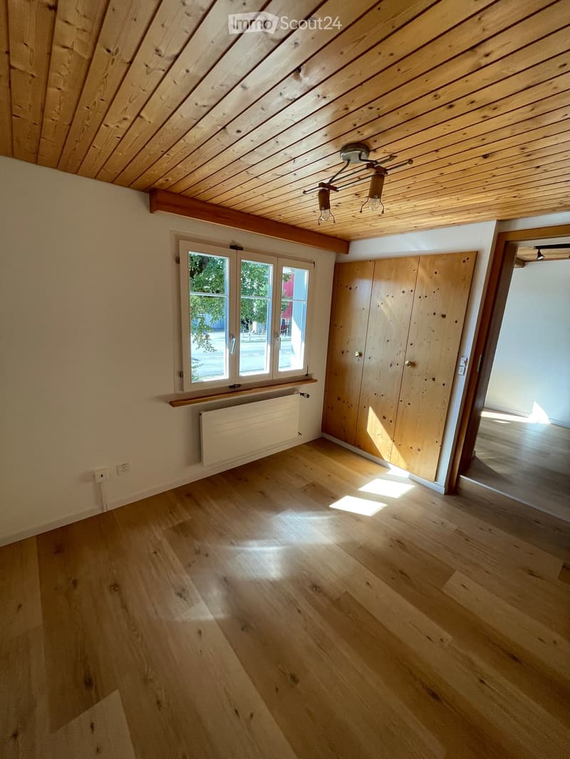 Empty room with wooden flooring, exposed wooden ceiling beams, and large windows allowing natural light. Features include a radiator, built-in wooden cabinetry, and a modern light fixture.
