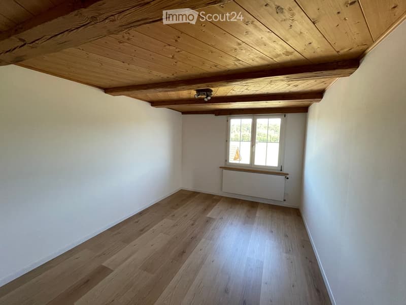 Empty room with wooden flooring, white walls, exposed wooden ceiling beams, and a single window with a radiator below it.