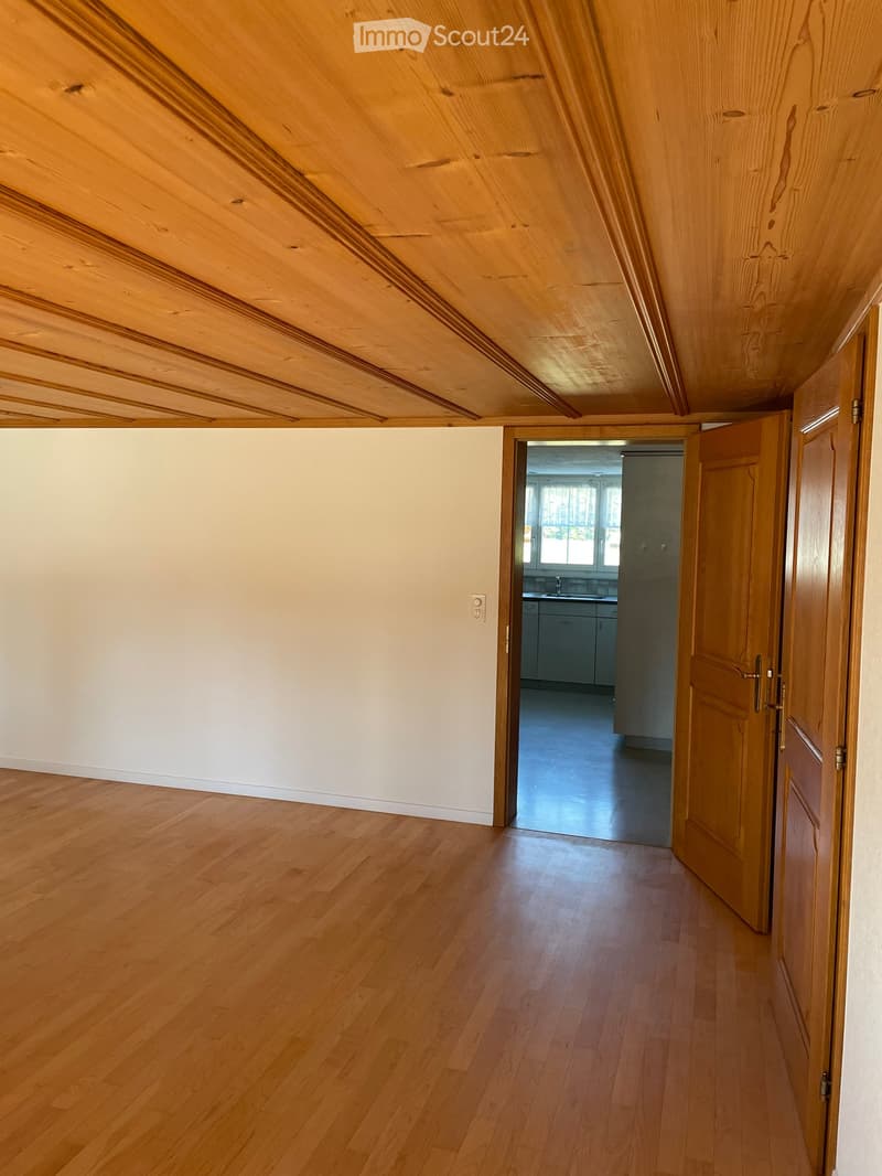 Empty hallway with wooden flooring and light-colored walls, leading to an adjacent room with a visible kitchen area.