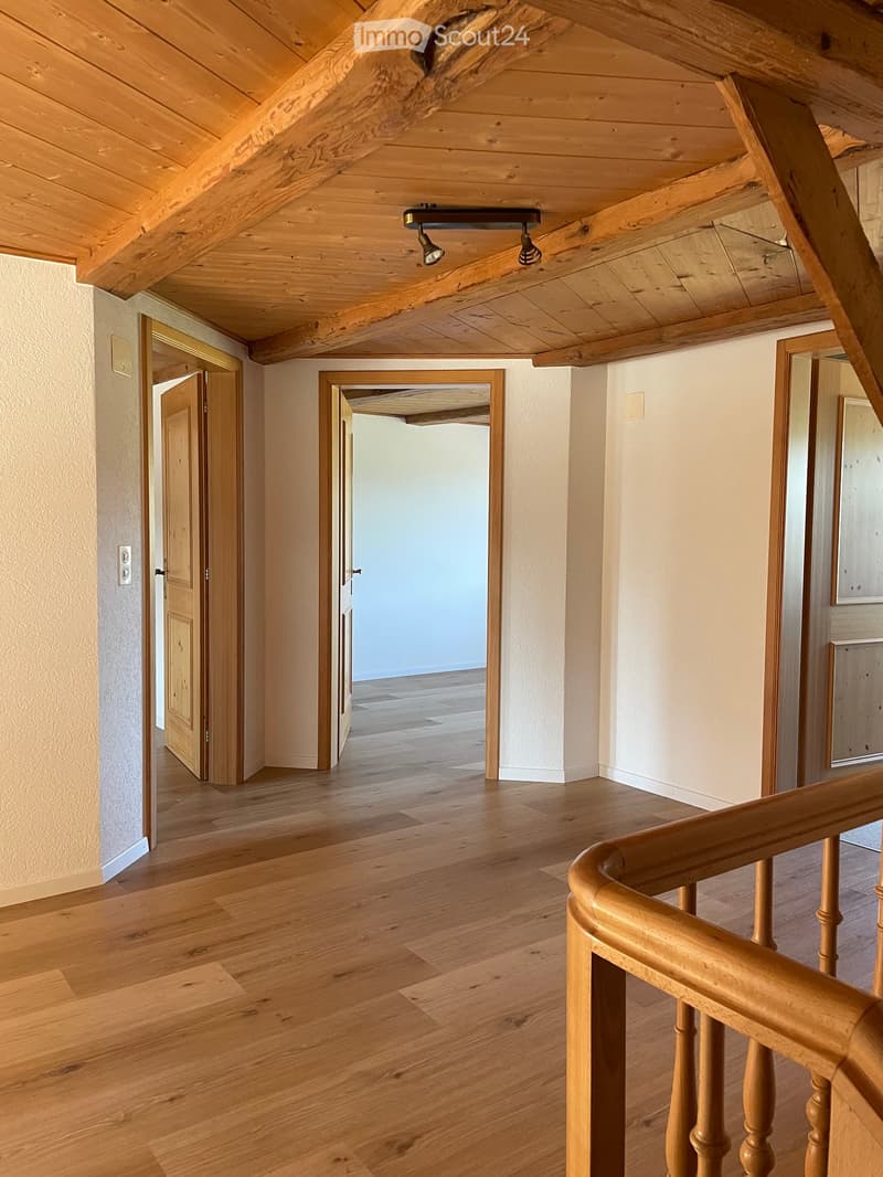 Wooden staircase with a curved railing and exposed wooden beams on the ceiling. The hallway has light-colored walls and wooden flooring, with several open doors leading to other rooms.