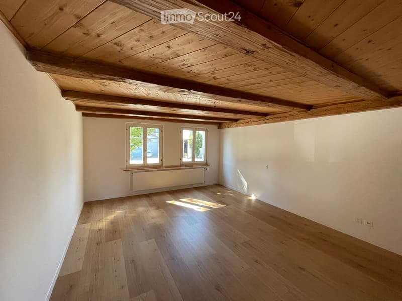 Empty room with wooden floor, white walls, exposed wooden ceiling beams, and two windows letting in natural light.
