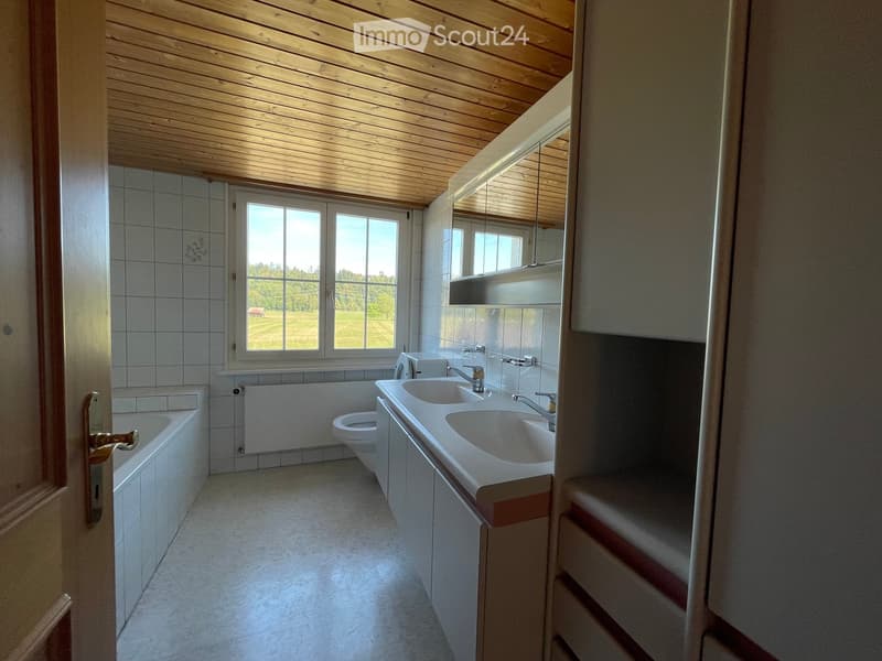 Bright bathroom with white tiled walls, a wooden ceiling, a large window offering a view of a green field, a bathtub, a toilet, a double sink, and a mirrored cabinet.