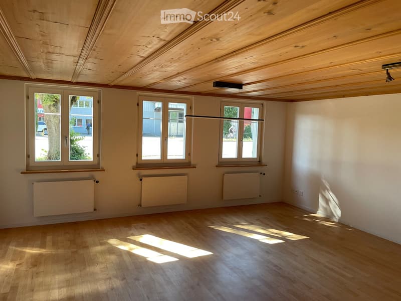 Empty room with wooden floor, white walls, exposed wooden ceiling, and multiple windows with radiators below. Natural light streams in, creating shadows on the floor.