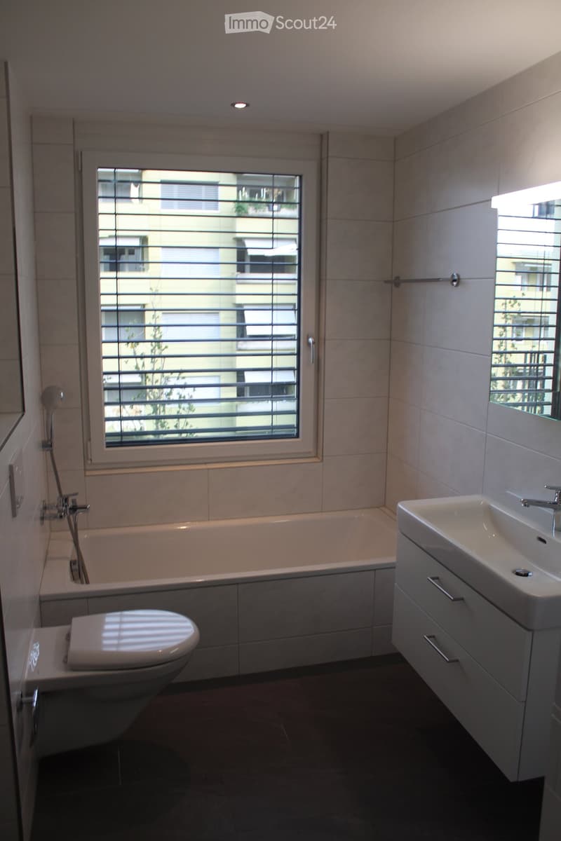 Modern bathroom with a bathtub, toilet, sink, and showerhead. Features large windows with blinds and a sleek, contemporary design.