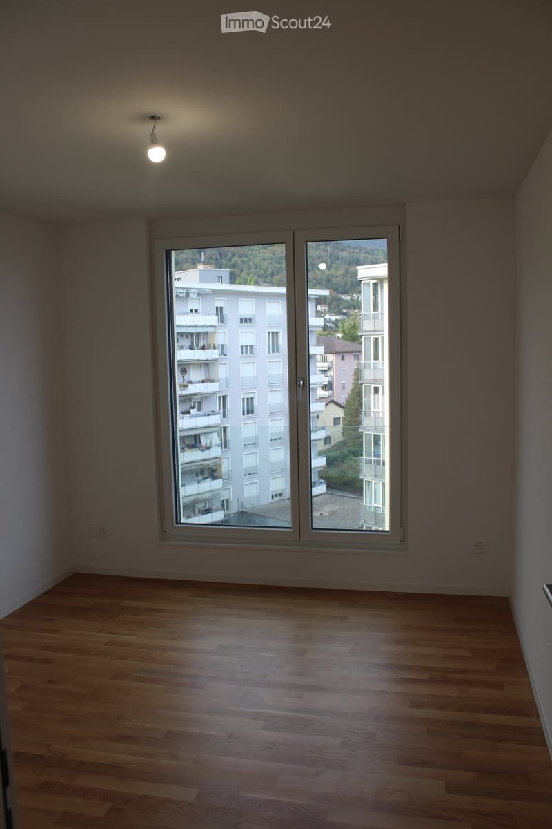 Empty room with wooden flooring and large windows offering a view of the cityscape and surrounding buildings.