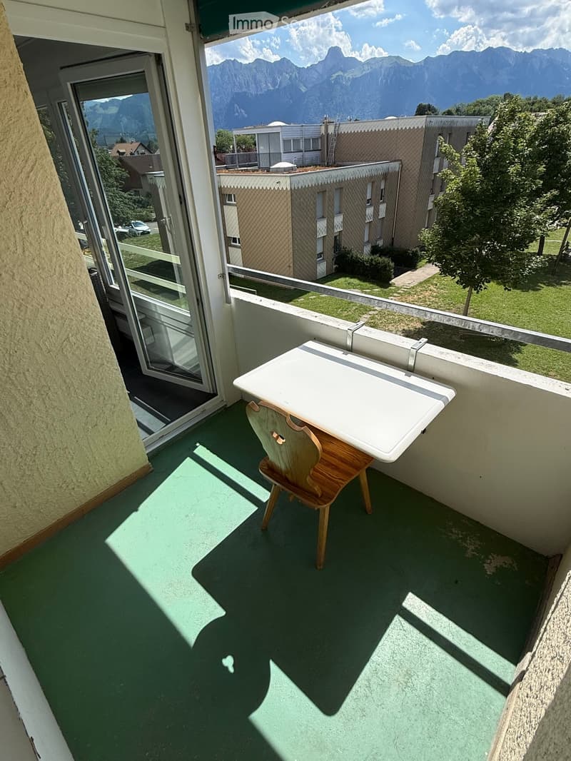 Balcony with green flooring, white table, and wooden chair, overlooking a scenic mountain view and neighboring buildings.