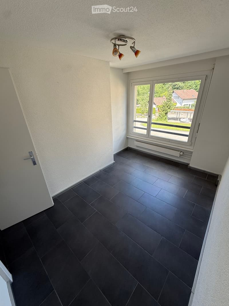 Empty room with dark tiled floor, white walls, large window with view of greenery, and modern ceiling light fixture.