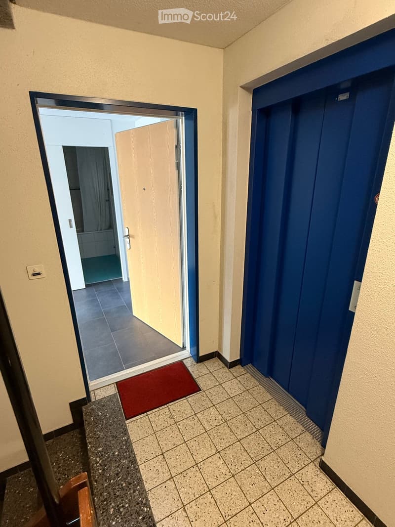 Hallway with tiled floor, beige walls, a red doormat, and an open door leading to another room.