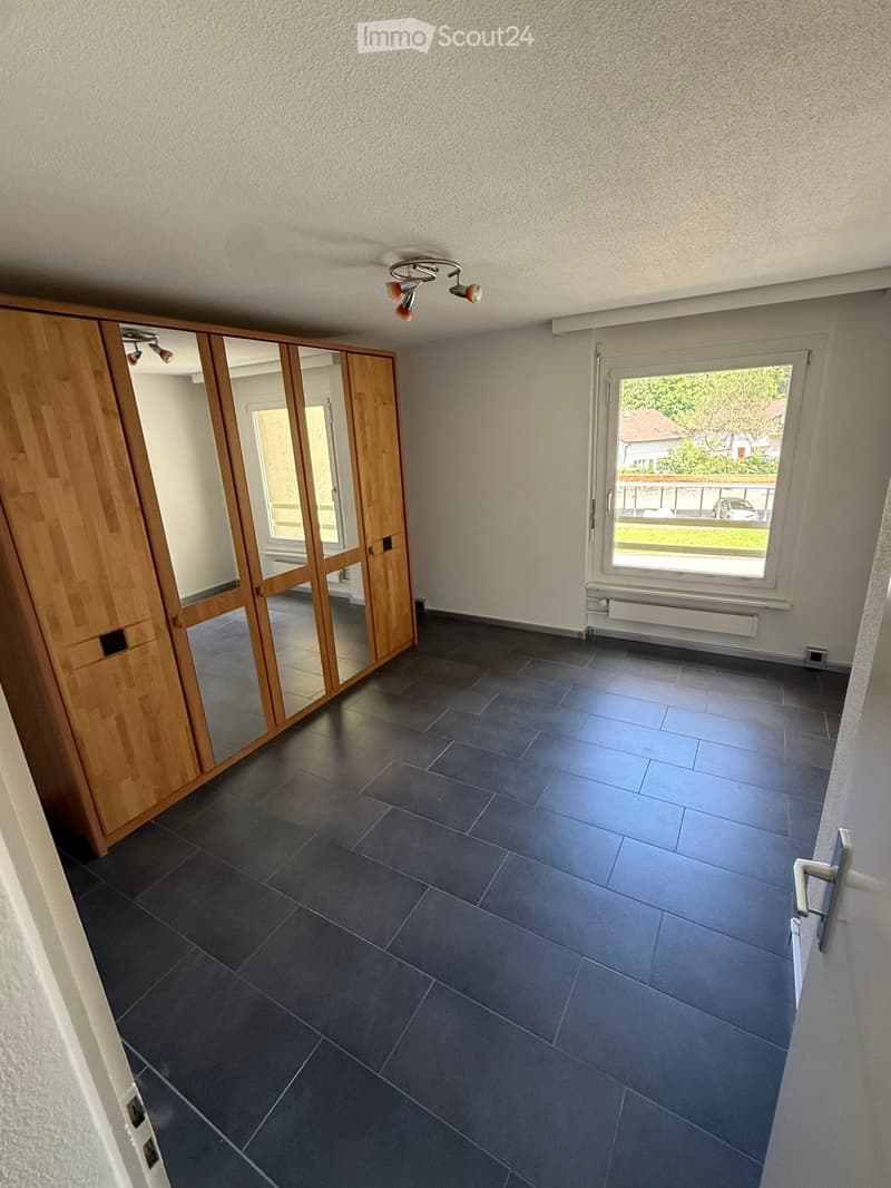 Empty room with dark grey tiled floor, light grey walls, large window with view of greenery, wooden wardrobe with mirrored doors, and ceiling light fixture.