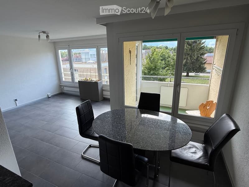 Dining area with a round table and four black chairs, large windows with a view of greenery, and a small balcony area with outdoor furniture.