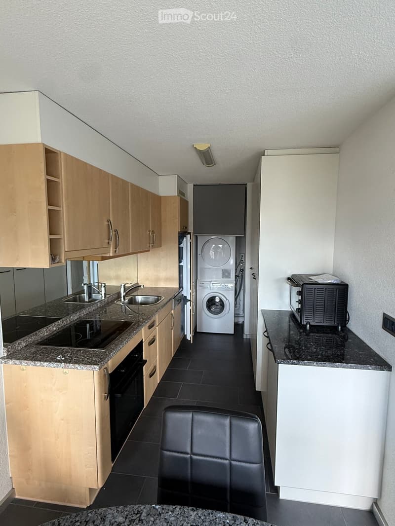 Modern kitchen with light wooden cabinets, granite countertops, stainless steel sink, washing machine, microwave, and a black leather chair.