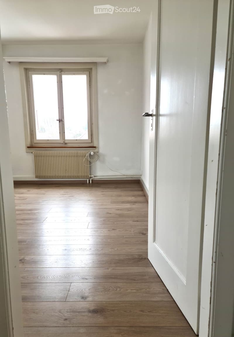 Empty room with wooden flooring, white walls, a window with a radiator below, and a door on the right side.