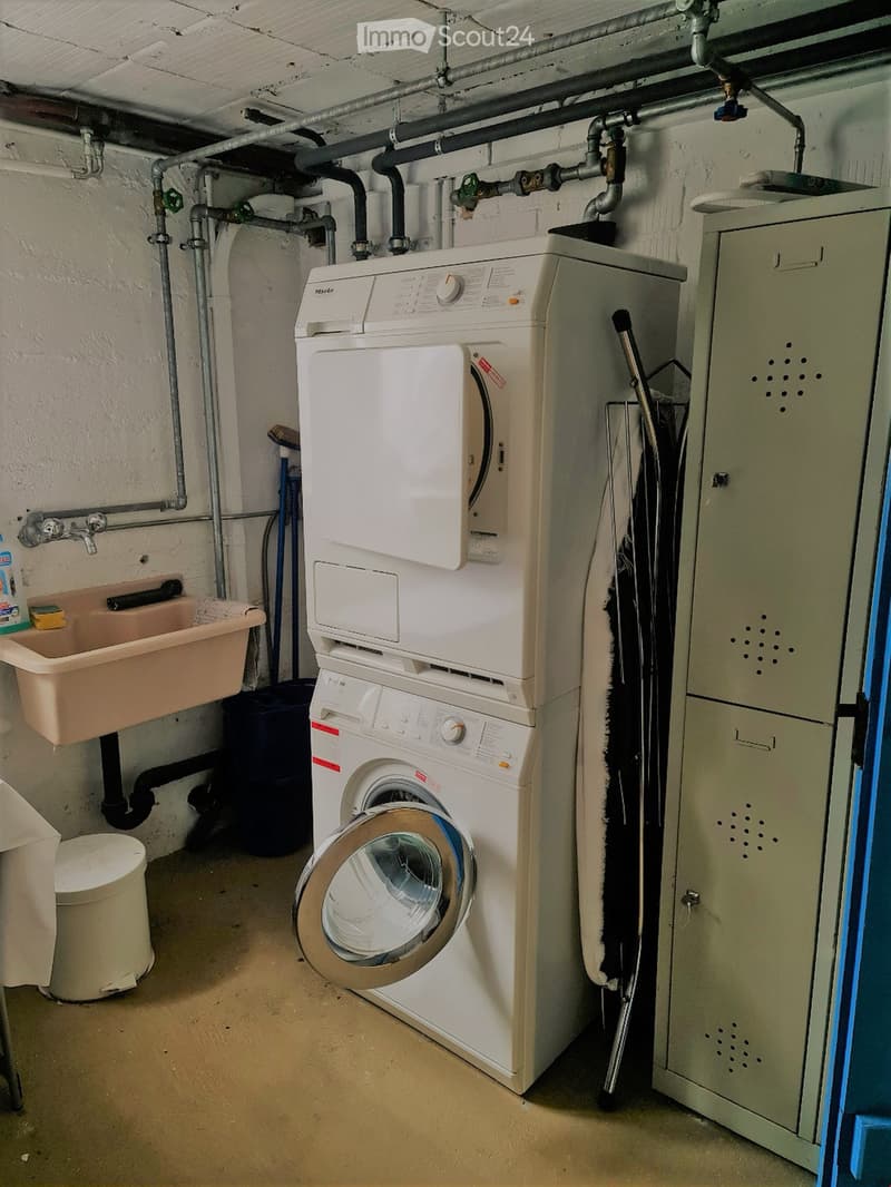 Utility room with a stacked washer and dryer, a sink, and a water heater. Exposed pipes and a concrete floor are visible.