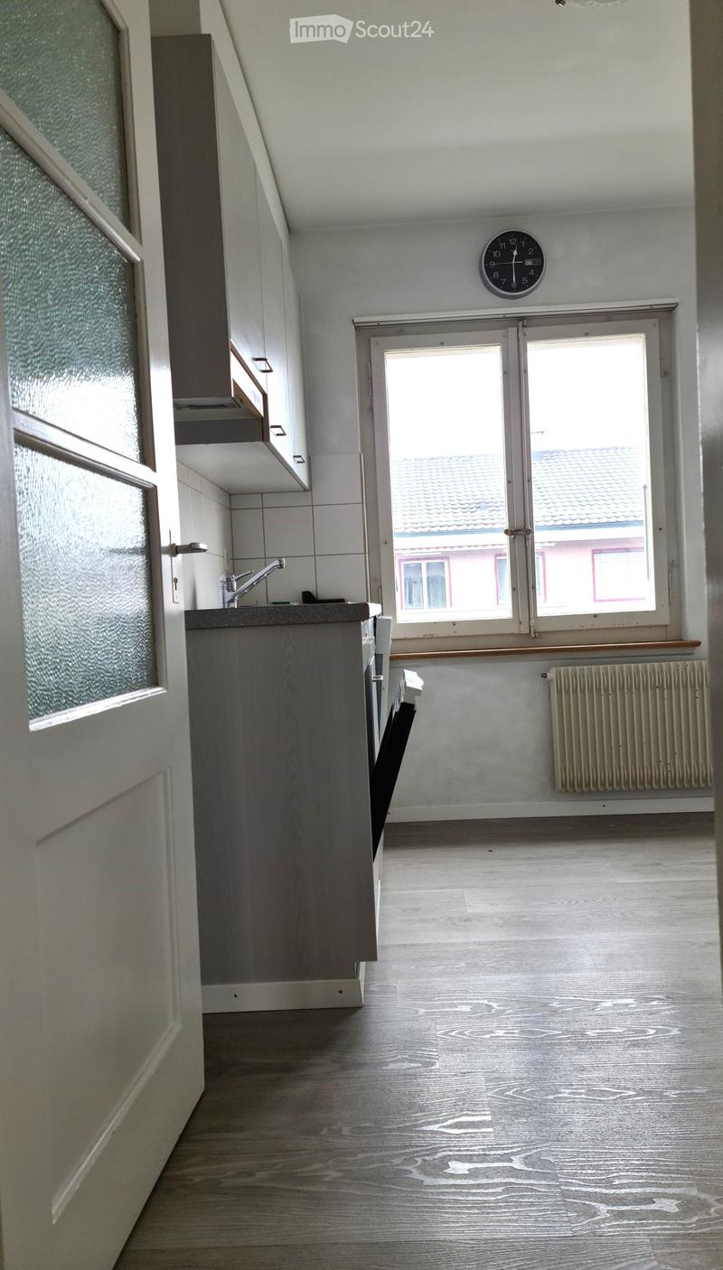 Empty kitchen with wooden flooring, white tiled walls, a sink, and a window overlooking a roof. No furniture or appliances are present.