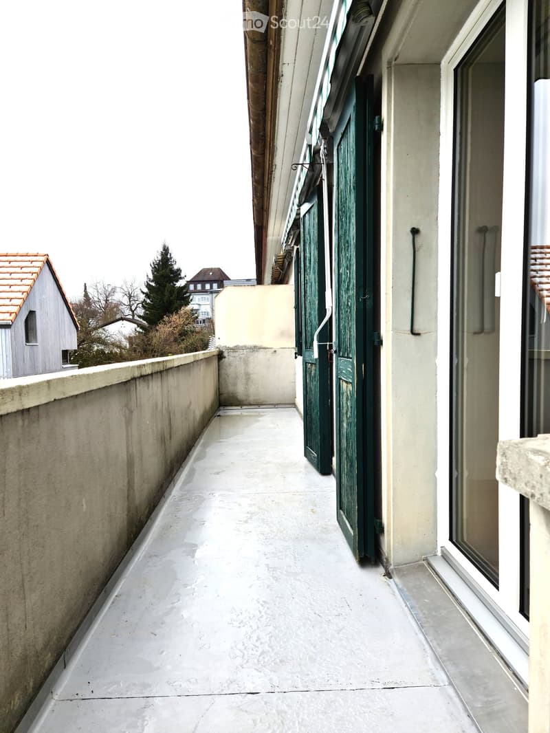 Narrow balcony with concrete floor, green door, and view of neighboring buildings and trees.
