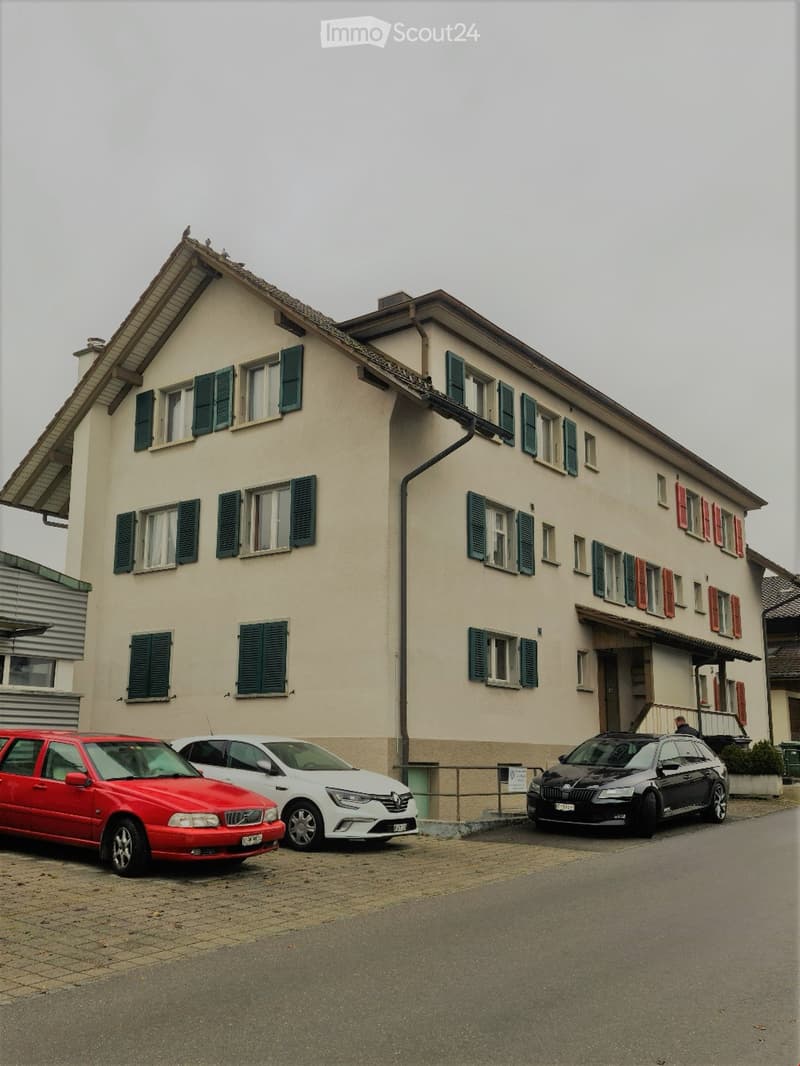 Two-story residential building with beige walls, green shutters, and multiple windows. Three cars are parked in front.