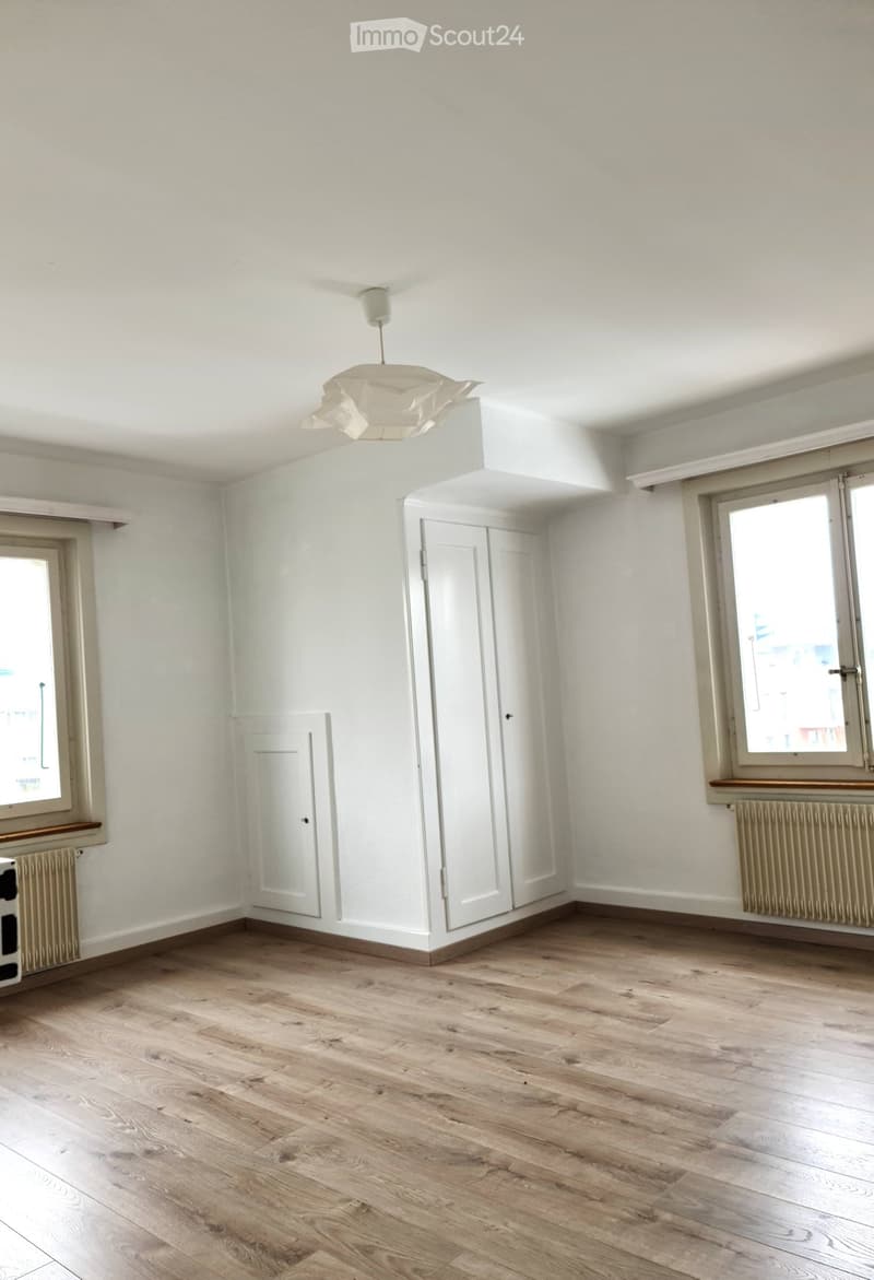 Empty room with light wooden flooring, white walls, and large windows. Features include a built-in closet, radiator, and a hanging light fixture.