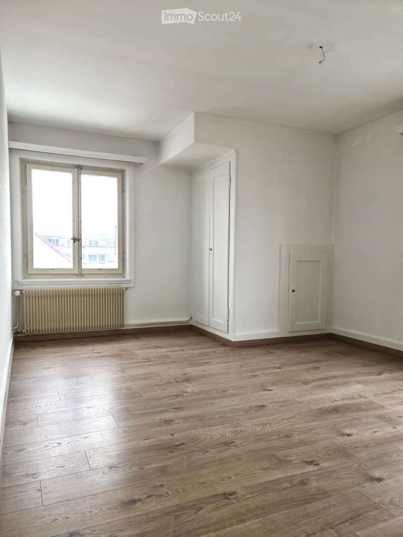 Empty room with light wooden flooring, white walls, a window with white frame, a radiator, and a closed door.