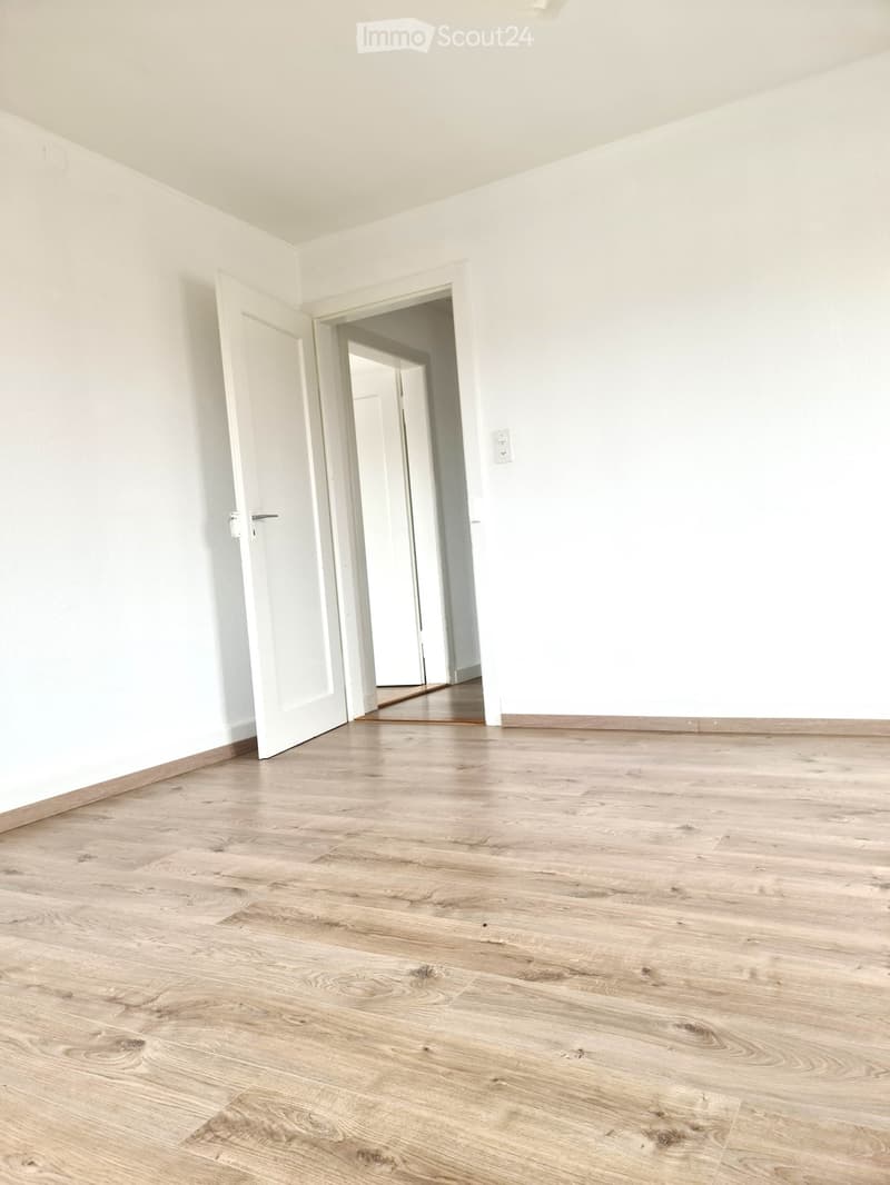 Empty room with light wooden flooring and white walls, featuring an open doorway leading to another room.