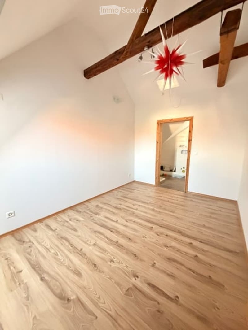 Empty room with wooden flooring, white walls, exposed wooden beams, and a doorway leading to another room.