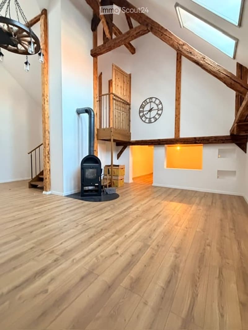 Open-plan space with wooden floors, exposed wooden beams, a staircase leading to an upper level, and a black wood-burning stove. The room is completely unfurnished.