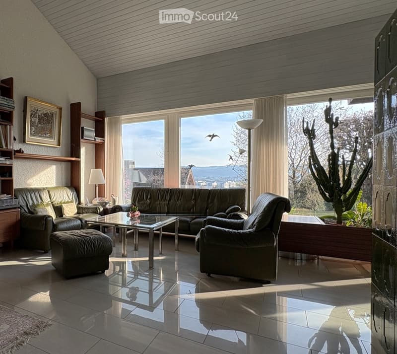 Modern living room with large windows offering a scenic view, featuring a dark green sofa, glass coffee table, armchairs, ottoman, floor lamp, table lamp, bookshelf, and picture frame.