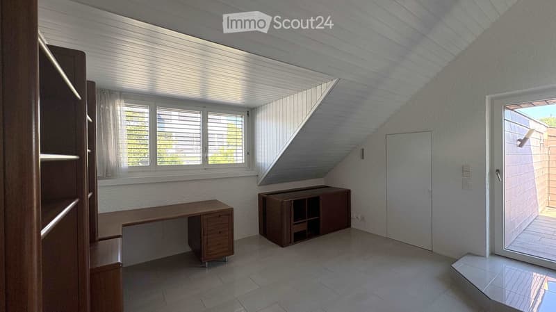 Bright, modern room with a slanted ceiling, large windows, and a door leading to a balcony. Contains a wooden desk, a cabinet, and a shelf.