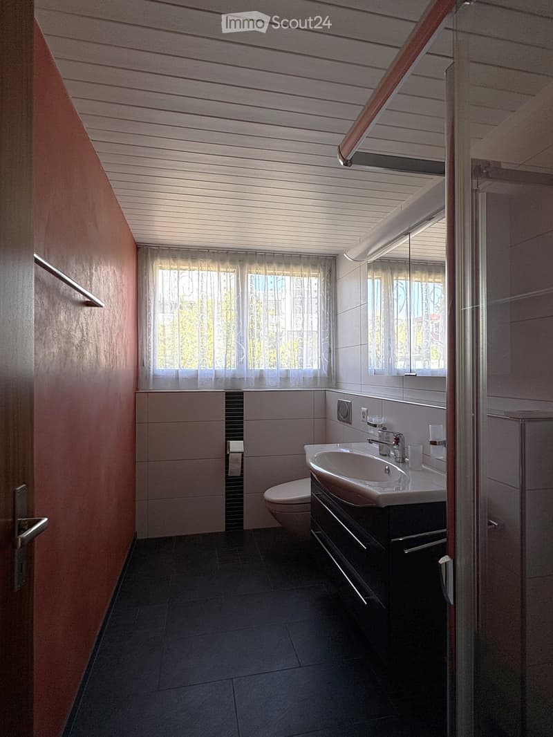 Modern bathroom with dark tiled floor, white sink, toilet, and mirrored cabinet. Features a window with sheer curtains and a glass shower enclosure.