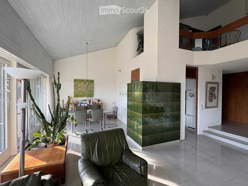 Modern living room with a green leather sofa, dining table with chairs, floor lamp, large window with view, and decorative cabinet.