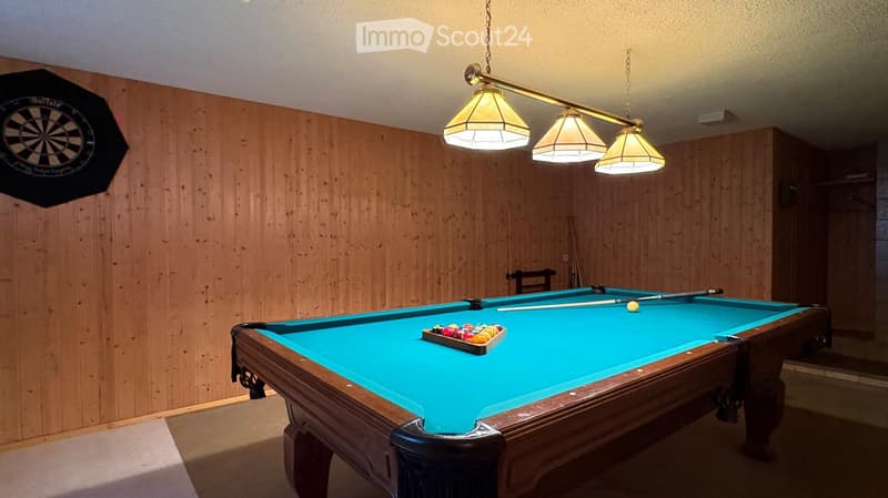 A room with wooden walls and a pool table, featuring a dartboard on the wall and pendant lights above the table.