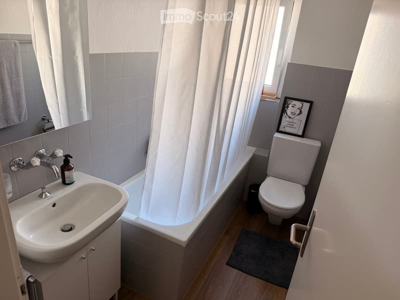 Modern bathroom with a white sink, chrome faucet, bathtub with a shower curtain, toilet, and a towel on the wall. A picture frame is placed on the toilet tank.