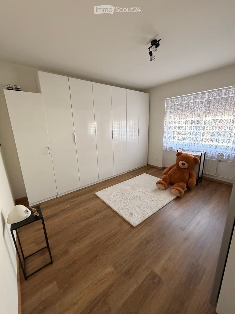 A furnished room with wooden flooring, white wardrobe, a small black stool with a hat and decorative item, a white rug, and a large teddy bear on the rug.