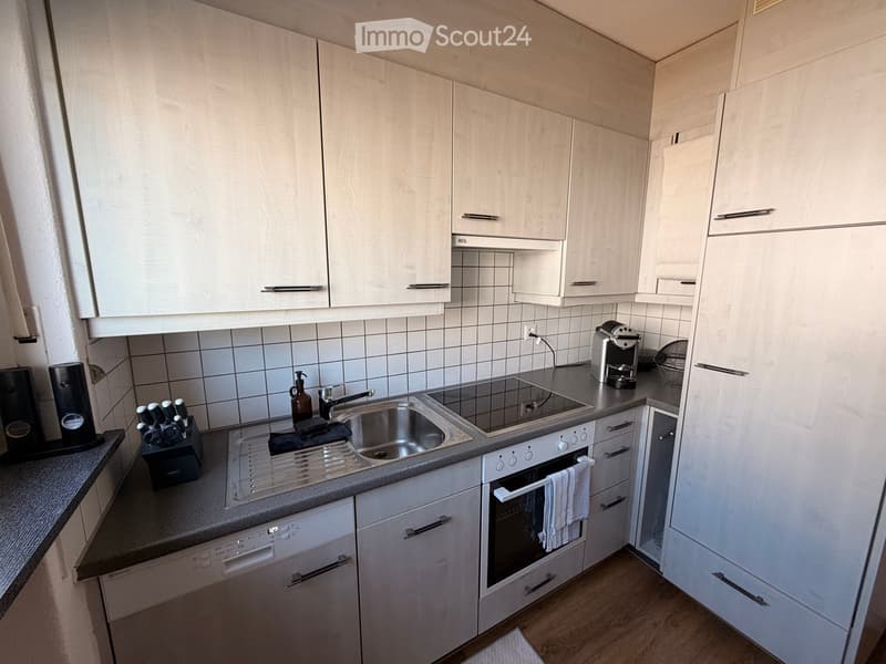 Modern kitchen with white cabinets, stainless steel appliances, and a grey countertop. Features include a sink, faucet, oven, dishwasher, coffee maker, and knife block.