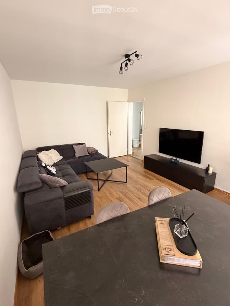 Modern living room with a grey sectional sofa, coffee table, TV stand, and a dining table with chairs. The room features wooden flooring and a sleek, contemporary design.